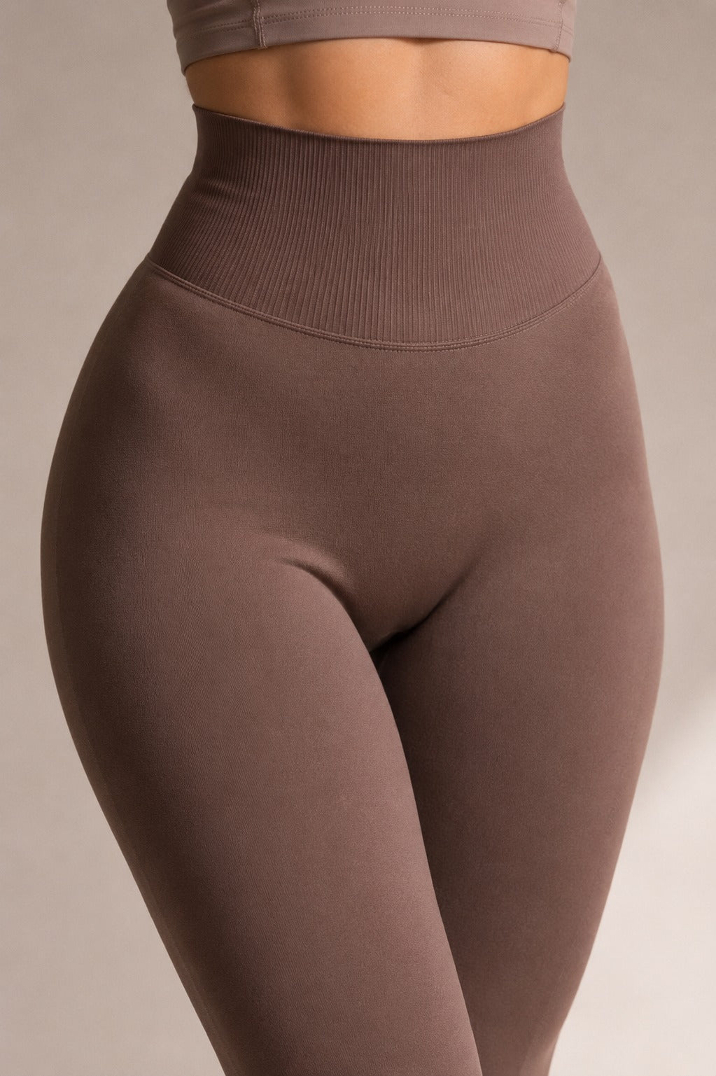 A close-up of a model wearing high-waisted, ribbed athletic leggings in a muted taupe-brown color, paired with a matching sports bra against a neutral studio background.